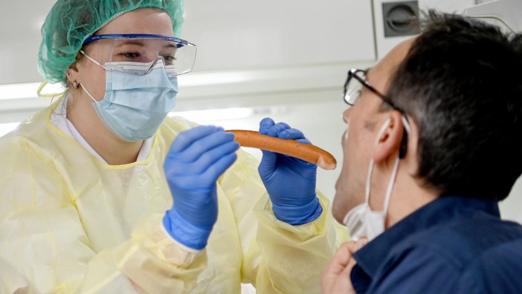 Sanitäterin schiebt einer Testperson ein Frankfurter Würstchen in den Rachen