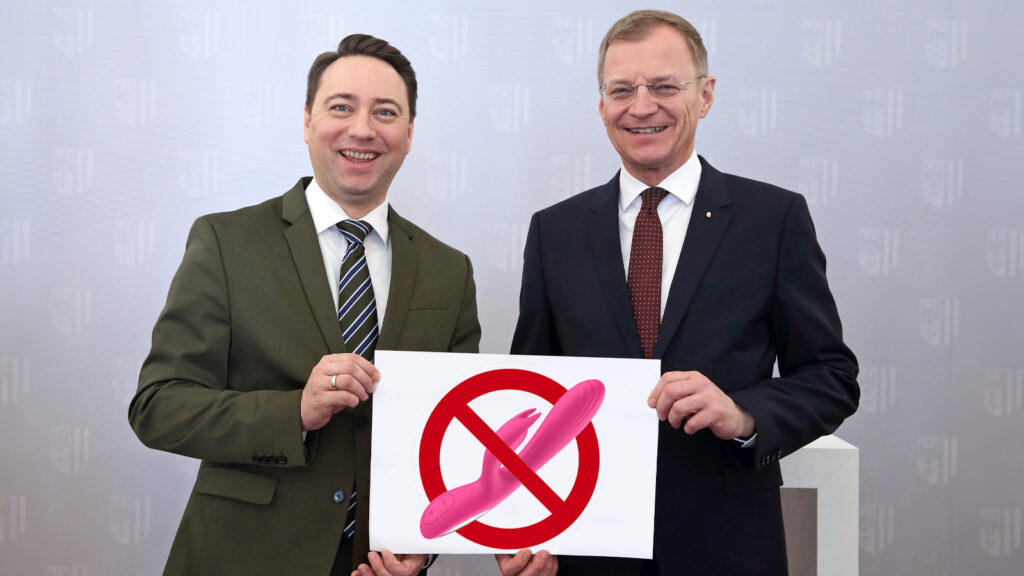 Manfred Haimbuchner und Thomas Stelzer halten eine Tafel mit einem durchgestrichenen Vibrator. Sie lächeln.