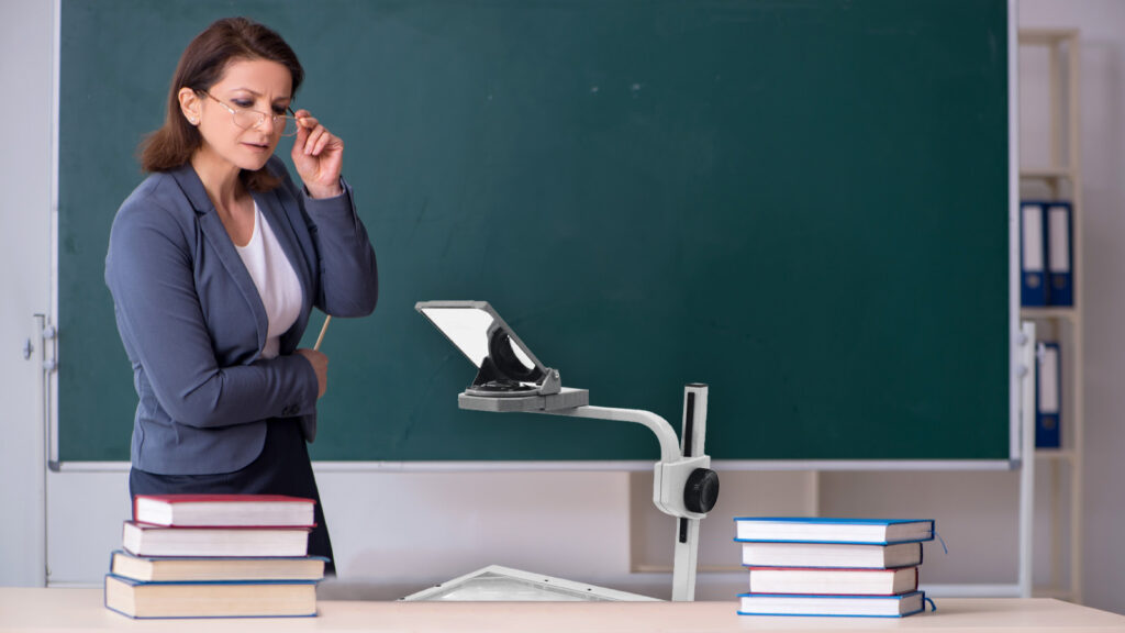 Overheadprojektor an einem Schultisch, neben ihm Bücher und die Lehrerin.