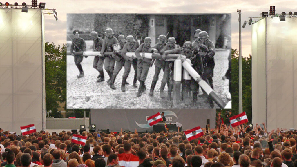 Eine großes Public Viewing, auf der Leiwand laufen Szenen aus dem 2. Weltkrieg.