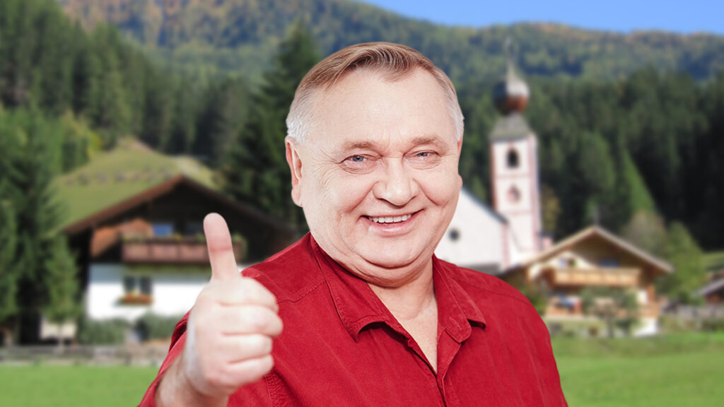 Ein Mann lächelt in die Kamera, dahinter ein kleines Dorf in den Alpen.
