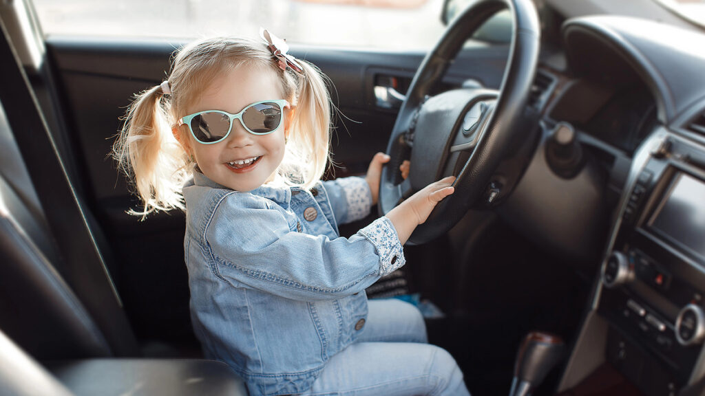 Das Bild zeigt ein kleines Mädchen mit Sonnenbrille am Steuer eines Autos.