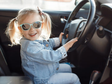 Das Bild zeigt ein kleines Mädchen mit Sonnenbrille am Steuer eines Autos.