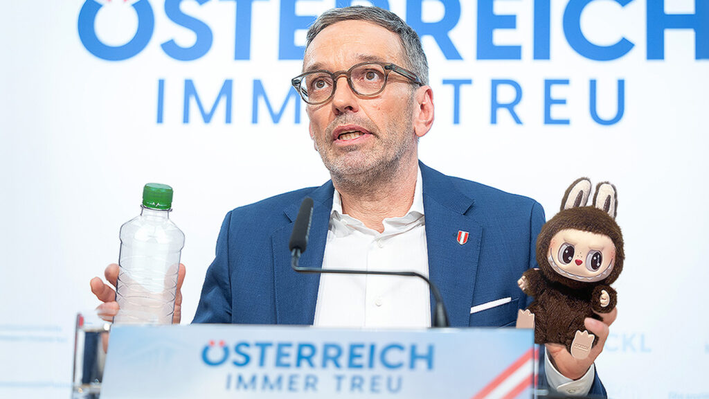 Das Bild zeigt Herbert Kickl bei einer Pressekonferenz, in einer Hand hält er eine Pfandflasche, in der anderen ein Labubu.