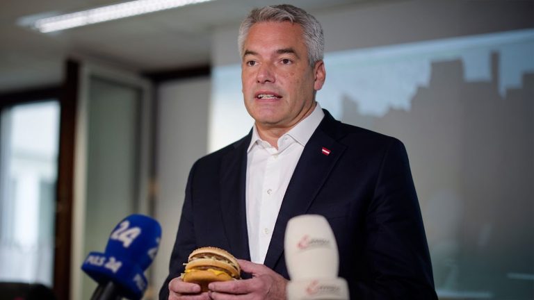 Das Bild zeigt Altbundeskanzler Nehammer mit einem Hamburger in Händen.