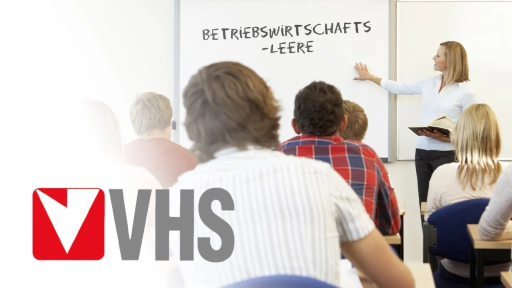 Das Bild zeigt das VHS Logo und eine Klasse voller Menschen.