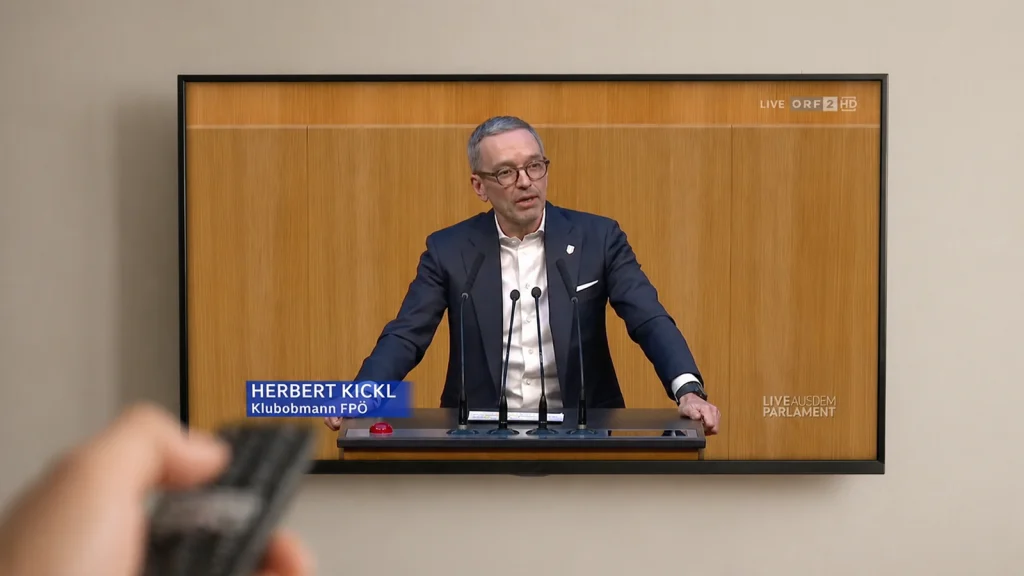 Das Bild zeigt eine Hand, die mit einer Fernbedienung einen Bildschirm bedient, auf dem Herbert Kickl zu sehen ist.