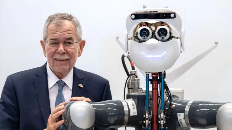 Das Bild zeigt einen lächelnden Bundespräsident Alexander Van der Bellen. Er lehnt sich an die Schulter eines Roboters.