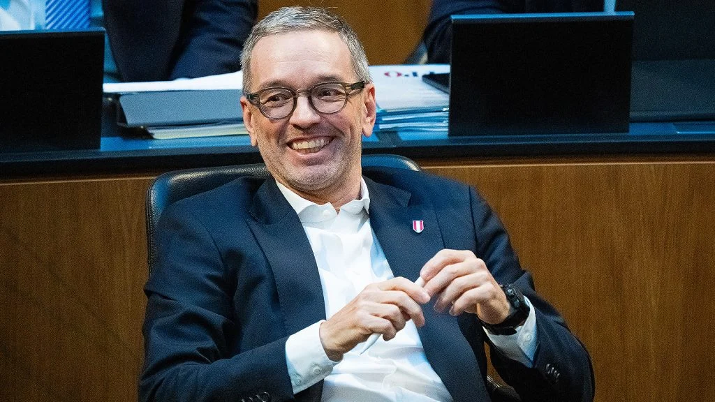 Das Bild zeigt einen lachenden Herbert Kickl, der zurückgelehnt im Parlament sitzt.